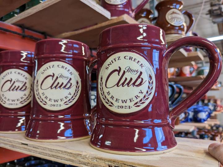 Red beer steins lined up on a plank of wood with with text "Snitz Creek Mug Club Brewery 2023".