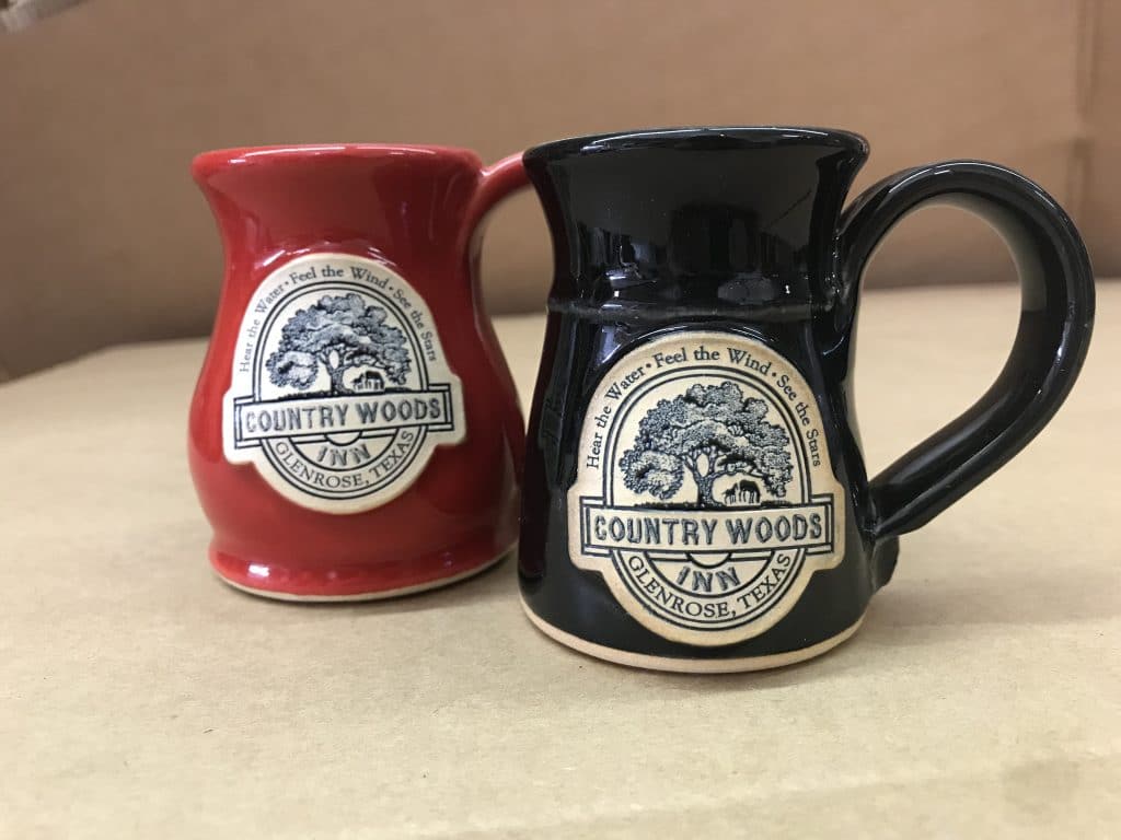 Red and black coffee mugs with logo medallions that say "Country Woods Inn Glenrose, Texas" and an illustration of a tree in the background.