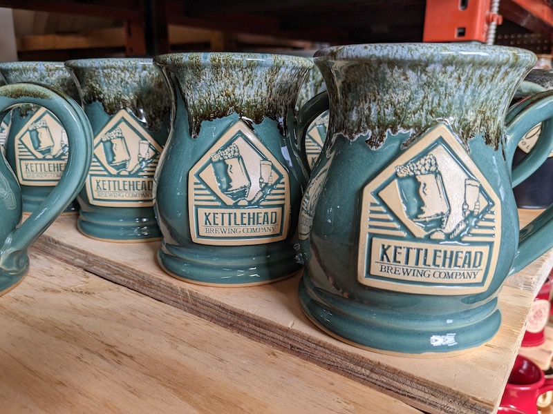 Green beer steins on a wood plank with an illustration and the text "kettlehead brewing" on a clay logo medallion.