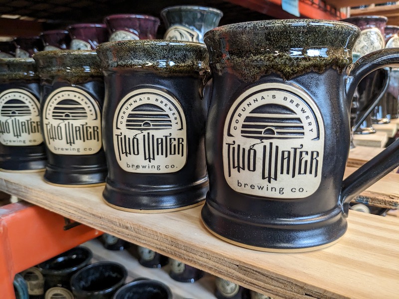 Black beer mugs on a wood plank with a clay logo medallion with the words "Two Waters Brewing".