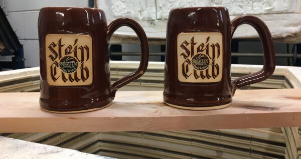 Two brown beer steins with logo stamps reading "Stein Club" sitting on top of a wood plank.