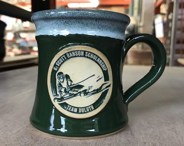 Green coffee mug with an image of a downhill skier and the text 'G.Scott Ranson Scholarship, Team Duluth'.