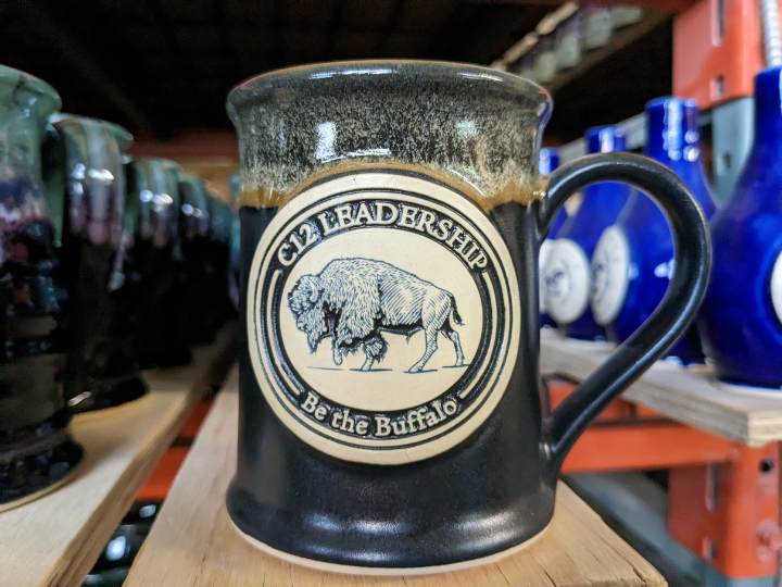 Black handmade coffee mug with clay medallion with illustration of a bison with the text "C12 Leadership. Be the buffalo."
