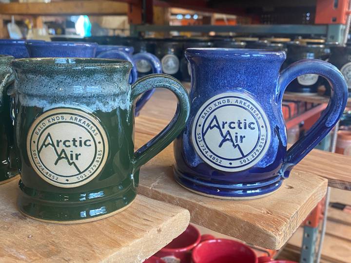 Green and blue coffee mugs with clay logo sitting on a brown wood board.