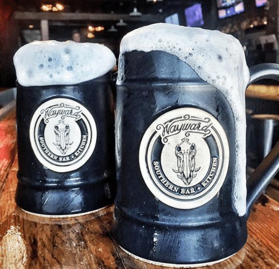 Two black beer steins with brewery logos on the front, filled with a froth on a bar counter.