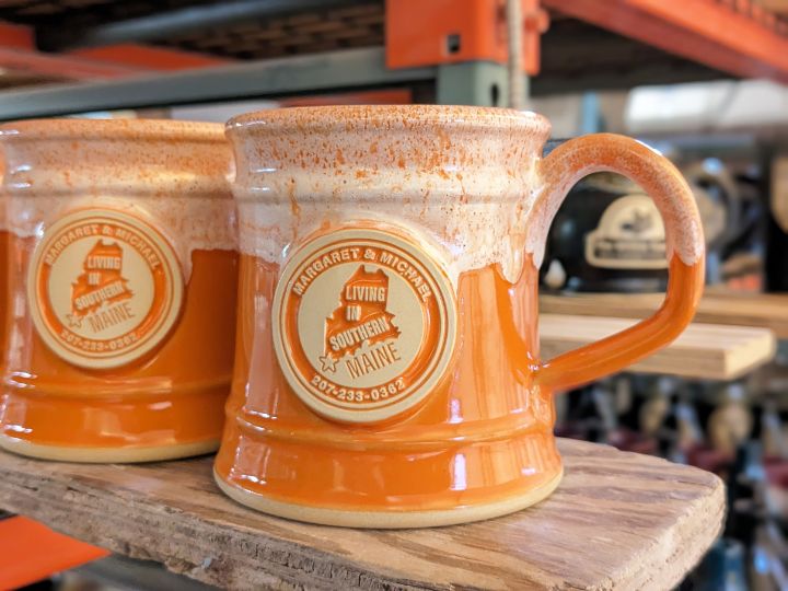 two orange and white mugs with the outline of the state of maine and text of two names, tagline and phone number