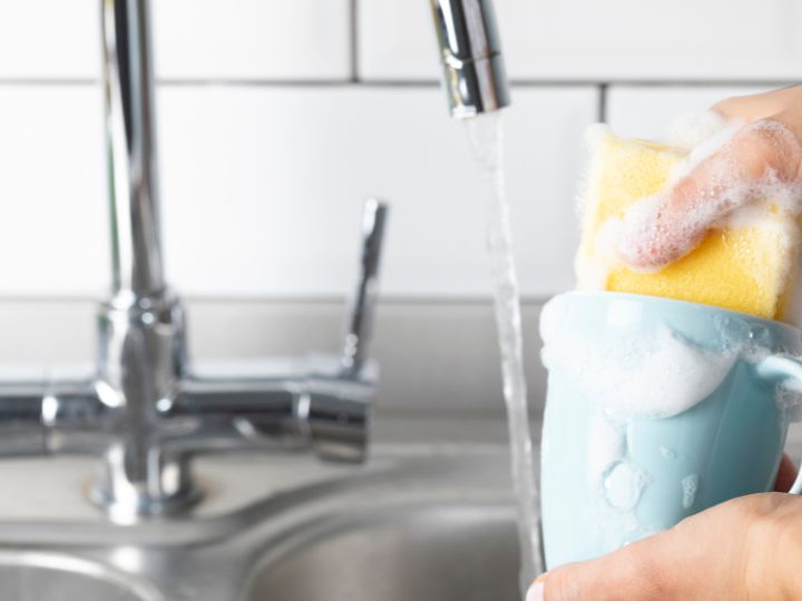 person cleaning a blue coffee mug under running water with soap and a sponge