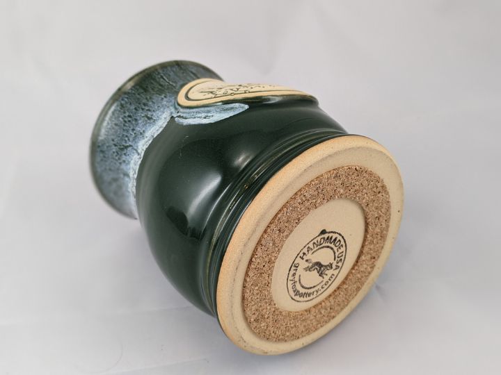 green coffee mug laying on its side with the bottom of the mug shown with a cork pad attached, label on the bottom of the mug reads 'handmade USA greyfoxpottery.com'