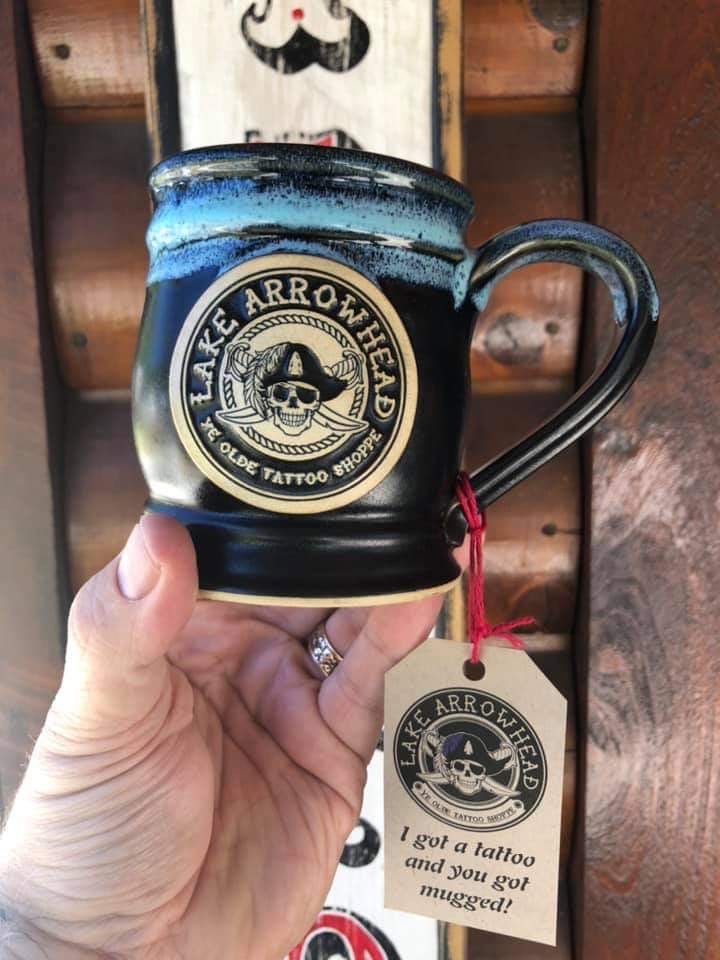 hand holding a black coffee mug with blue glazed rim. the coffee mug has a round clay medallion logo of a pirate tattoo shop