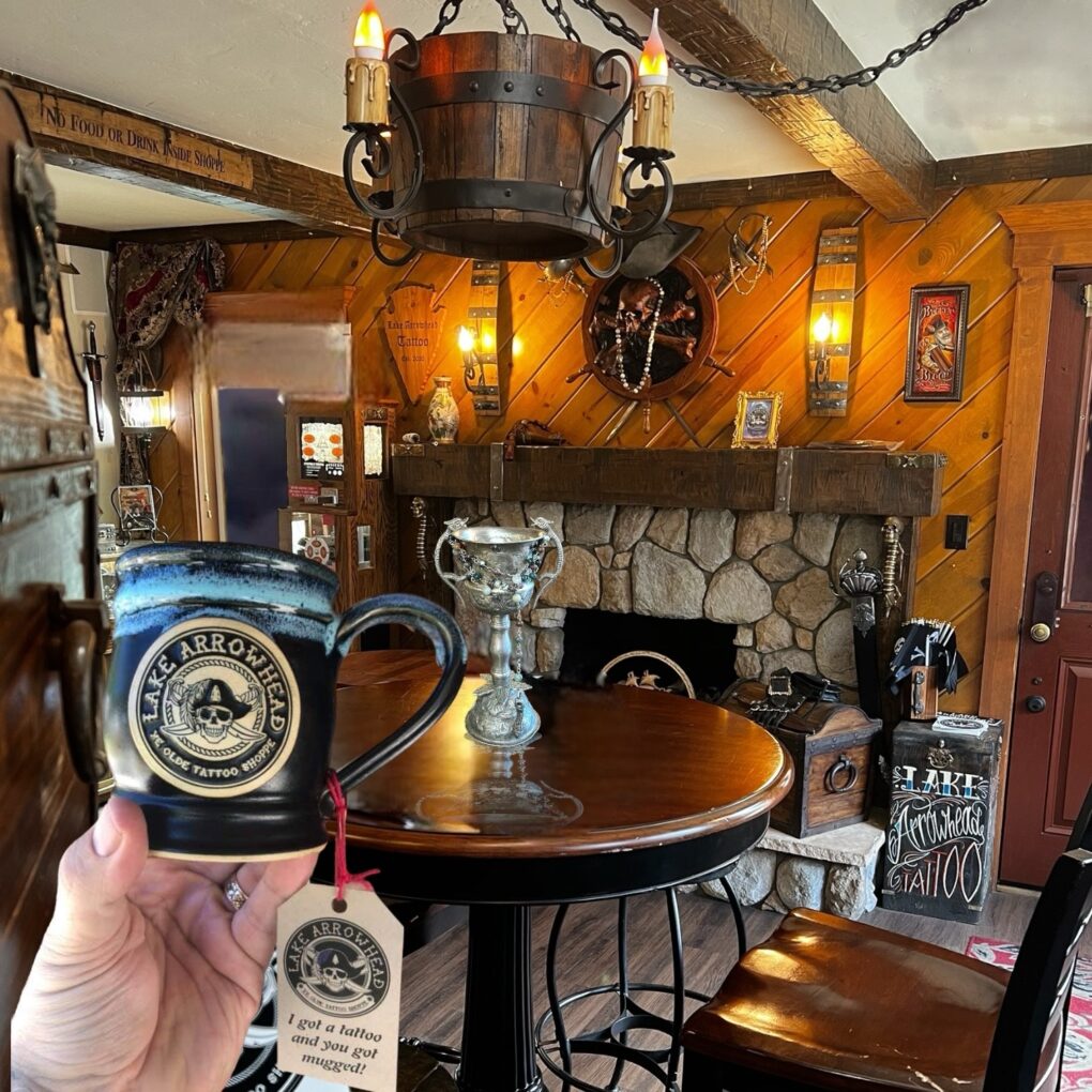 hand holding a black coffee mug with blue glazed rim. the coffee mug has a round clay medallion logo of a pirate tattoo shop. in the background is a room with a fireplace and old world decor