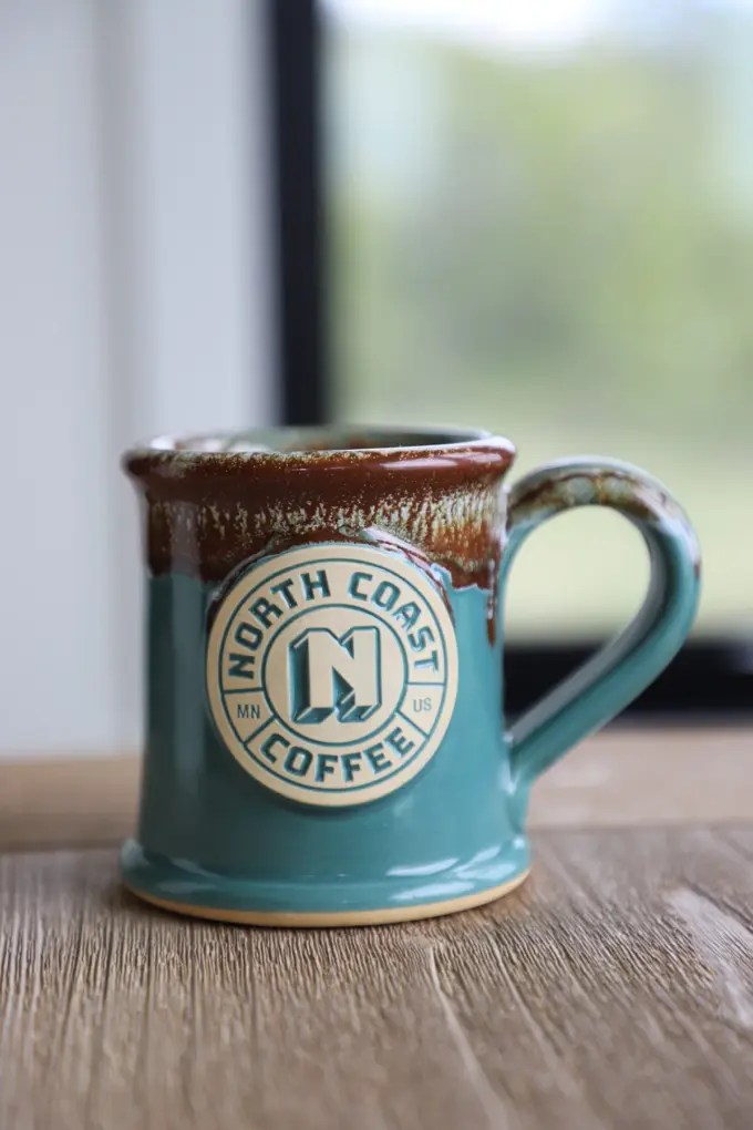 logo coffee mug in teal and brown glaze, sitting on a table near a window