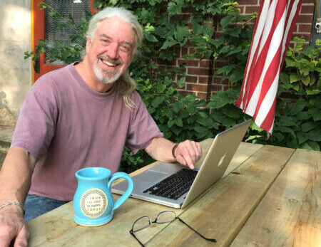 man sitting in front of a laptop computer with a coffee mug and eyeglasses next to him on a table.