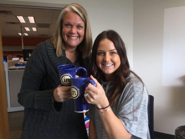 two women standing together holding blue coffee mugs with their company logo on them.