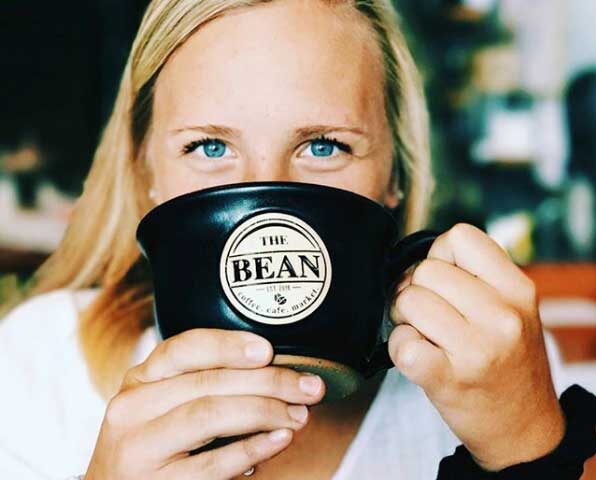 a woman holding a large black coffee mug with a logo for the bean coffee company up to her face