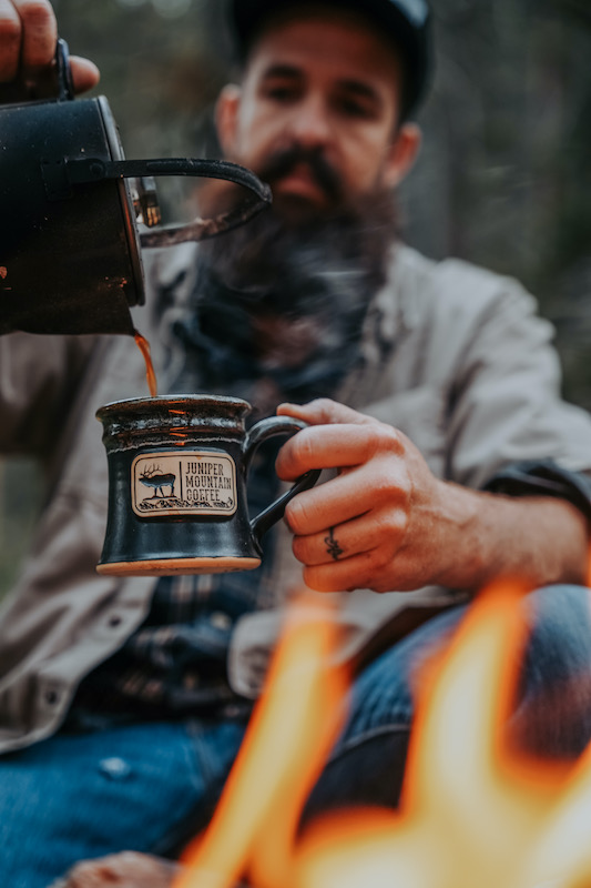 man pouring coffee into a mug sitting next to a campfire<br />
